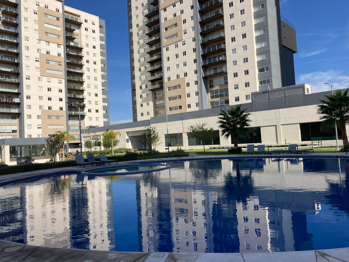 A large swimming pool is featured in the courtyard, reflecting the nearby buildings and sky. Surrounding the pool are lounge chairs and well-maintained grassy areas, providing an inviting space for relaxation and leisure.
