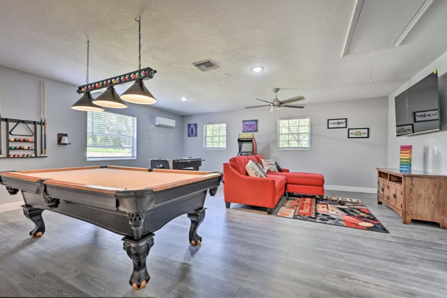 A game room features a sleek pool table and a cozy seating area with a bright orange couch. A wall-mounted Smart TV is visible along with decorative artwork and a ceiling fan. Natural light enters through two windows, enhancing the playful atmosphere.