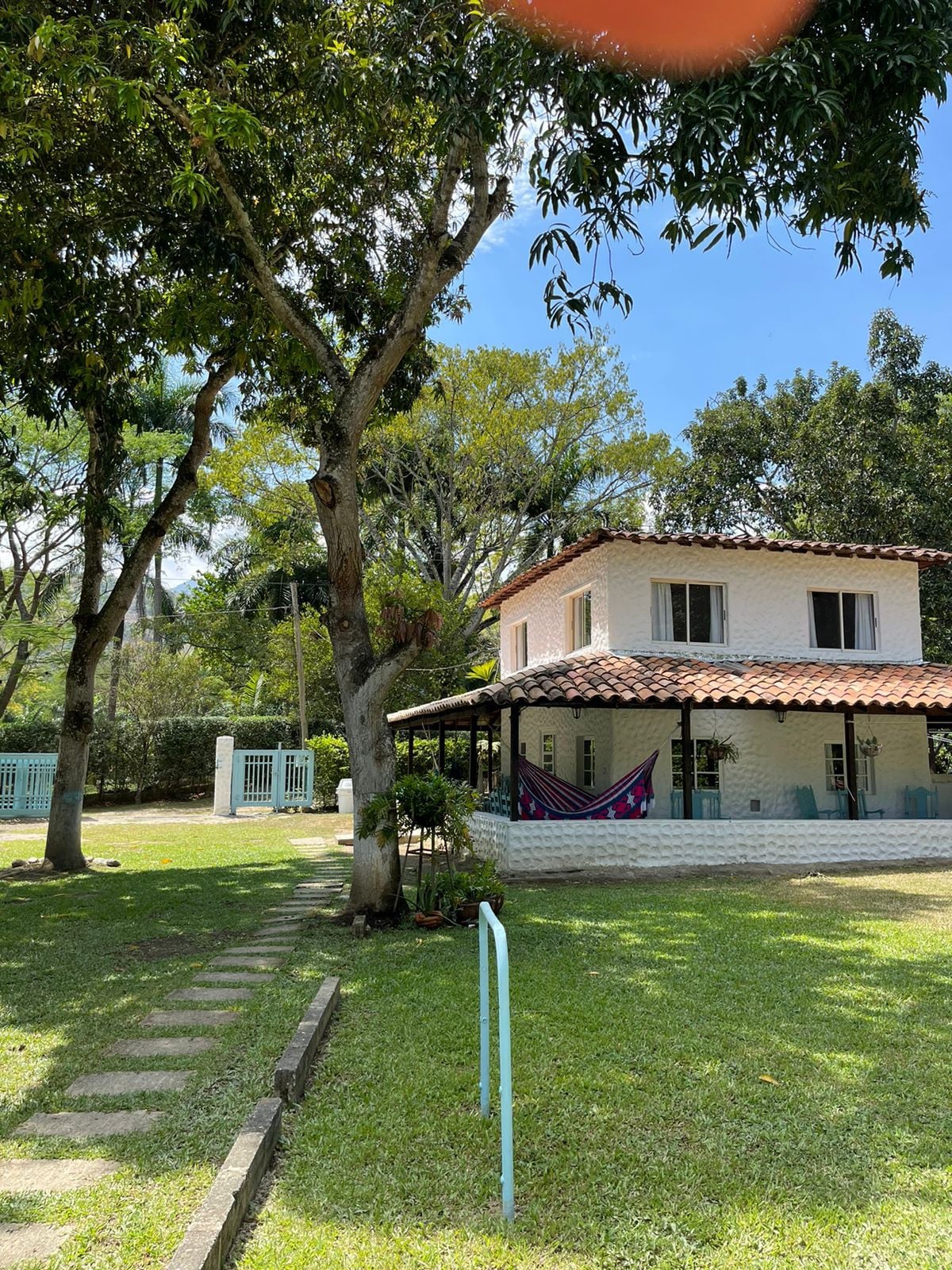The two-story finca is surrounded by lush greenery, featuring a covered porch with a hammock. Pathways of stone lead to the entrance, complemented by large trees offering shade. The house showcases a traditional tile roof and bright walls, creating a welcoming outdoor atmosphere.