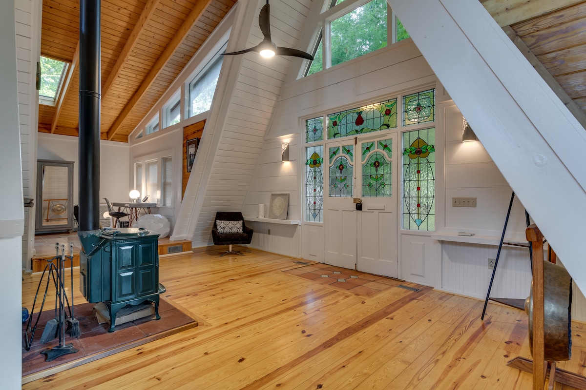A spacious living area is highlighted by vaulted wood ceilings and large windows that provide ample natural light. Stained glass entry doors contribute color while a wood-burning stove sits centrally on a brick platform. Light wood flooring complements the overall design.