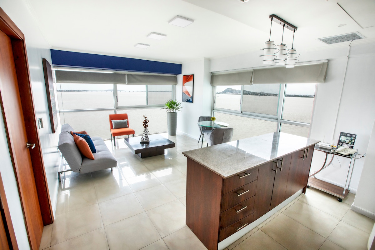 A bright living area features large windows providing views of the outside. A grey sofa is paired with an orange accent chair, and a wooden coffee table centers the space. The kitchen area includes a smooth countertop and stylish cabinetry.