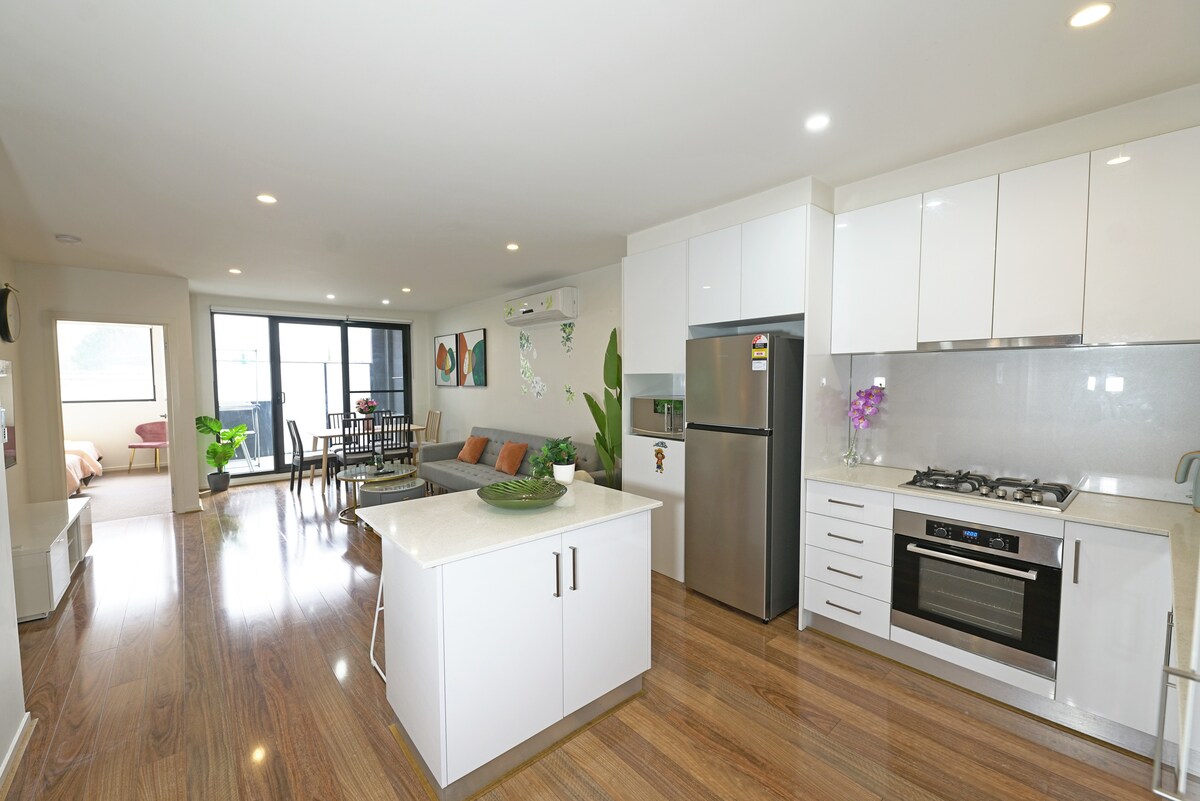 An open-plan living area features a modern kitchen with sleek white cabinetry and stainless steel appliances, including a gas stove and refrigerator. A dining table is set for six, complemented by a comfortable seating area with colorful cushions. Natural light floods in from large glass doors leading to a balcony.