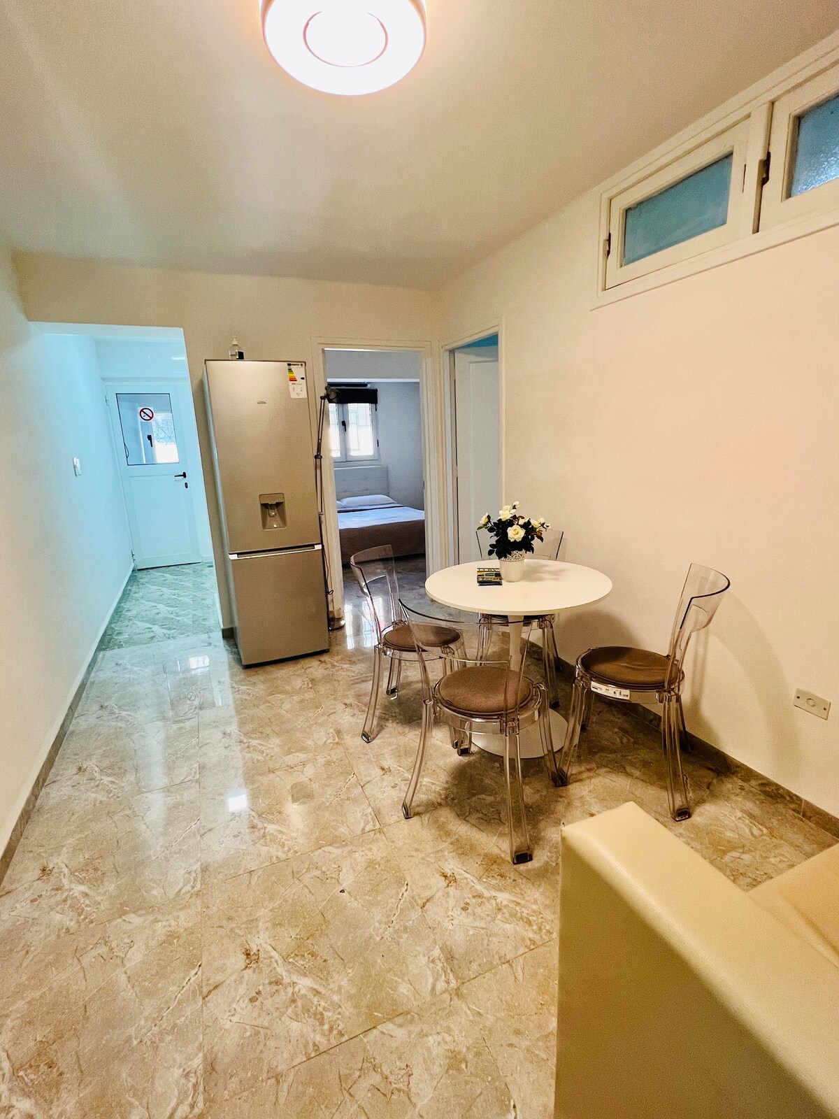 A compact kitchen area features a round dining table with transparent chairs, set against a backdrop of light-colored walls and marble flooring. A silver refrigerator is positioned adjacent to the table. Doorways visible in the background lead to additional rooms, including a softly lit bedroom.