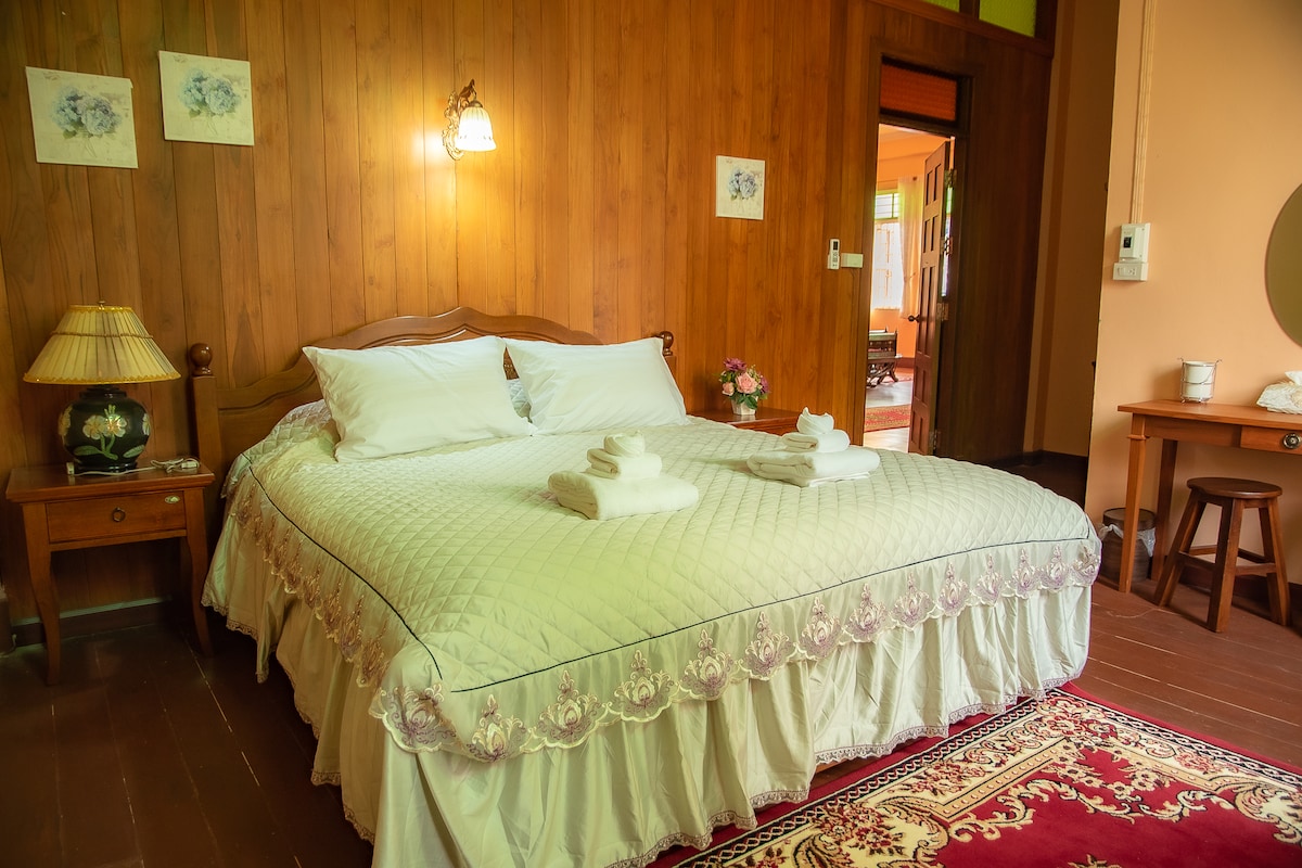 A double bed is positioned at the center of the room, complemented by a classic wooden headboard. Soft white linens cover the bed, adorned with neatly arranged towels. A warm wooden wall sets a cozy ambiance, while a bedside lamp and small table enhance the overall comfort.