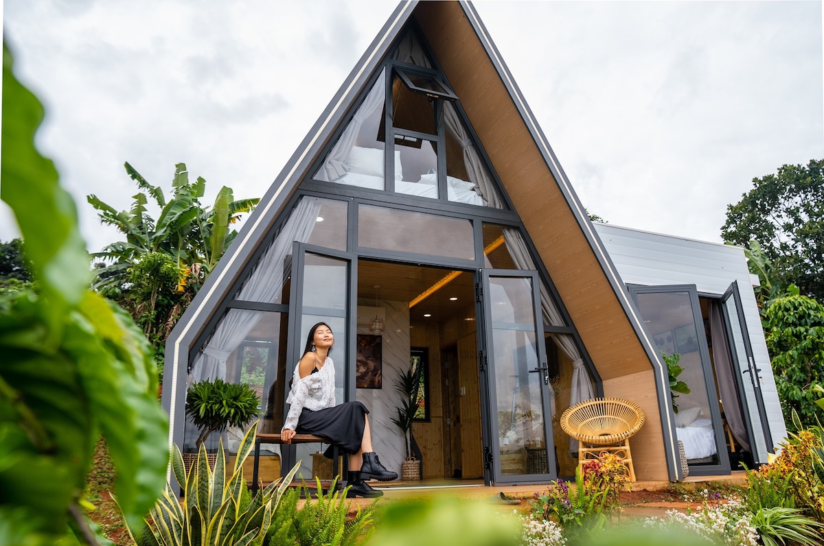 A classic A-frame home is displayed, featuring a full wall of windows that invite natural light. The entrance is framed by lush greenery and vibrant plants. A seating area is positioned outside, offering a relaxing space amidst a serene landscape.