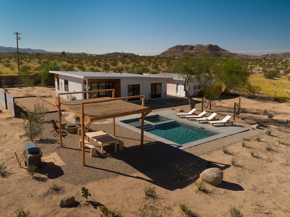 An aerial view captures Casa Bella's modern structure surrounded by desert landscape. The in-ground pool and spa are visible, along with sun loungers set on a deck. A shaded outdoor dining area can be seen, framed by minimal desert foliage and mountain views in the background.