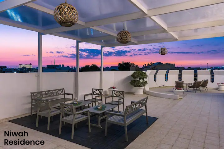A shared rooftop terrace features modern outdoor furniture, including a glass-top table and six chairs, arranged for relaxation. The area offers a view of a twilight sky painted in hues of purple and orange, with decorative hanging lanterns adding warmth.