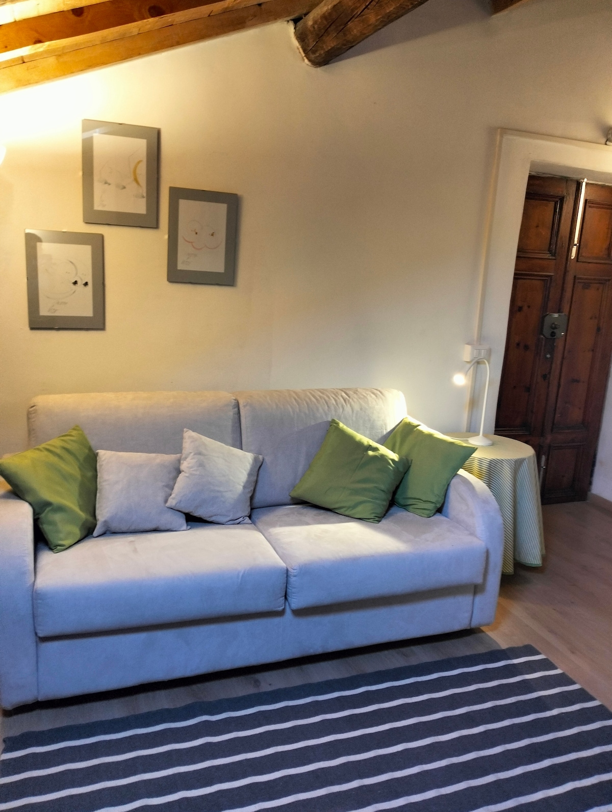 A light-filled living area features a beige sofa adorned with several decorative pillows in shades of green. Above the sofa, framed artwork adds a touch of character. A small side table is positioned beside the sofa, and wooden beams can be seen on the ceiling.