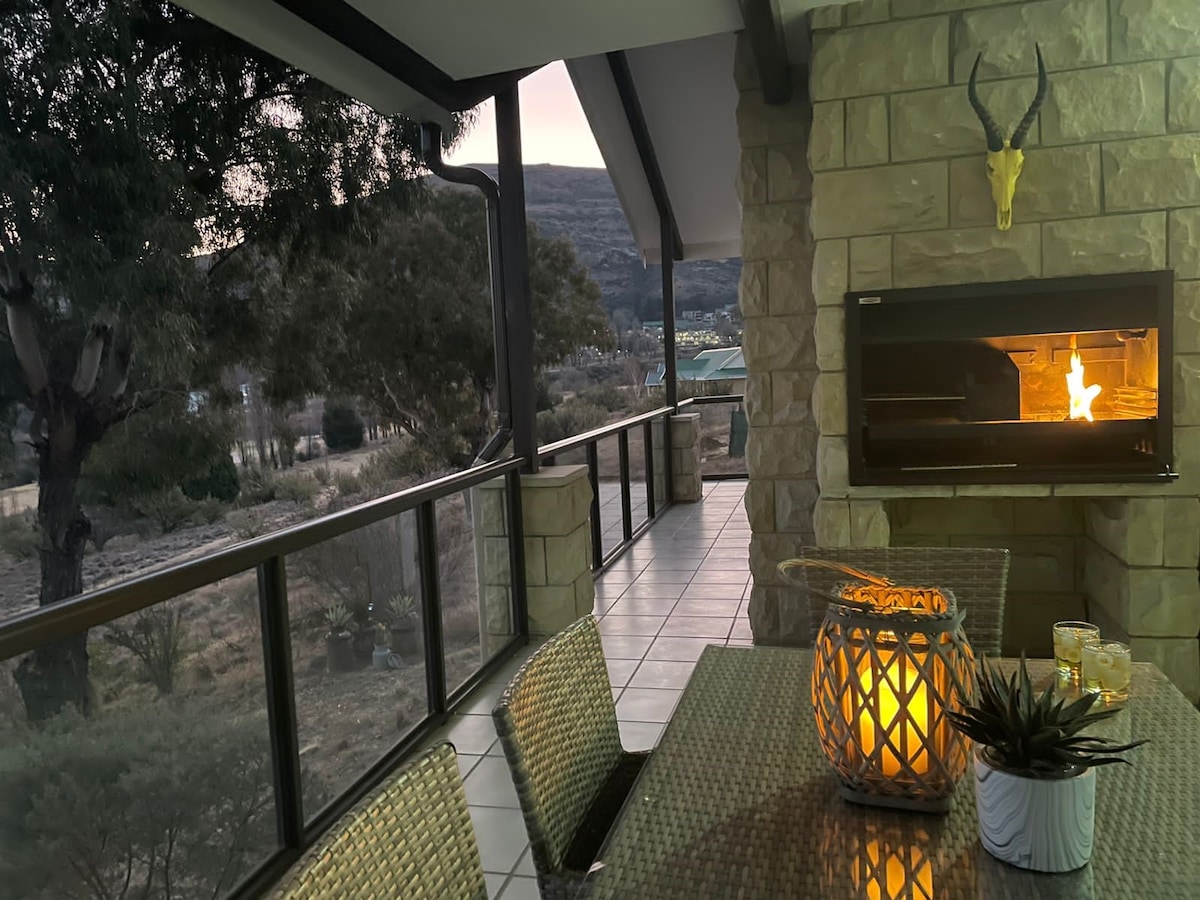 A large patio area is seen, featuring a built-in braai and a six-seater dining set. Soft lighting emanates from a decorative lantern, and the surrounding landscape includes trees and mountains, creating a tranquil outdoor atmosphere.