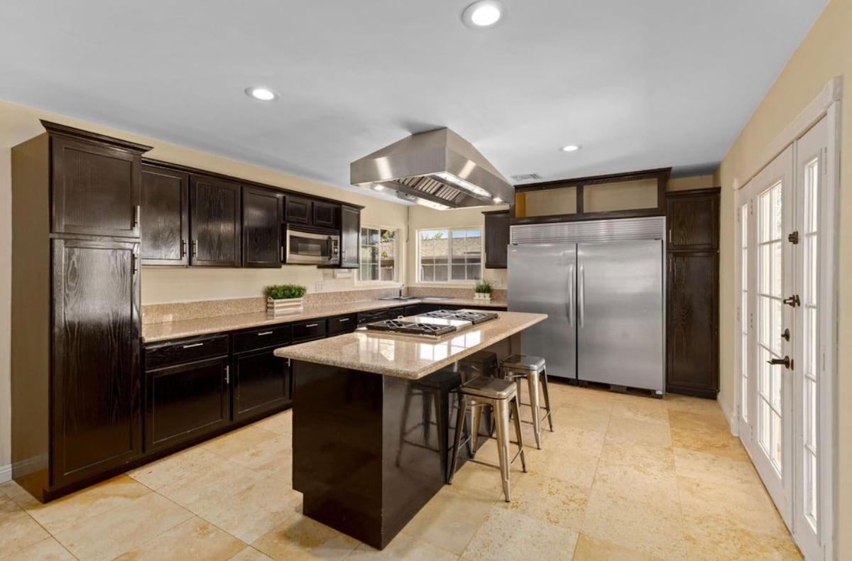 The modern kitchen features dark cabinetry and an expansive island with a gas cooktop. A stainless-steel refrigerator and oven are noted, complemented by stone countertops. Natural light enters through the glass doors, providing a view of the outdoor area.