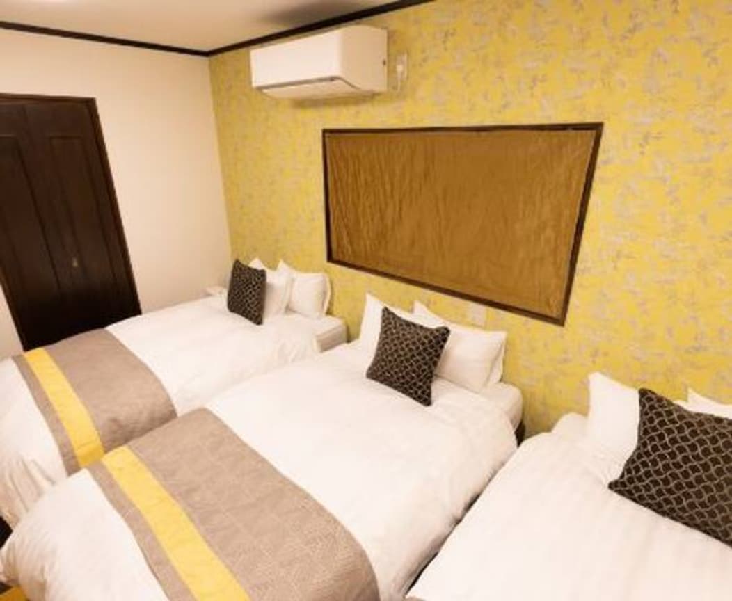 A well-appointed bedroom features three spacious beds with crisp white linens and decorative pillows. The yellow patterned wallpaper adds a vibrant touch to the room, while an air conditioning unit ensures a comfortable environment.