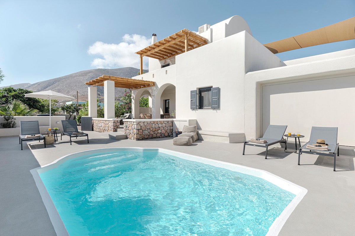 A private villa features a refreshing swimming pool surrounded by sun loungers and a solid stone outdoor bar. The structure boasts light-colored walls and an appealing pergola shading a seating area, with a scenic mountain backdrop visible beneath a clear blue sky.