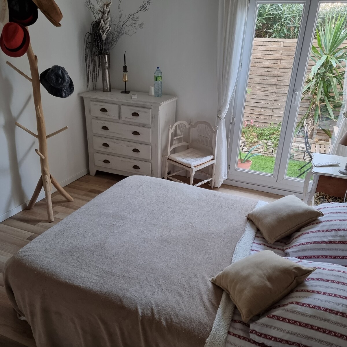 A bright bedroom features a spacious bed dressed in neutral tones. A light wooden dresser and a small chair provide functional decor. Large glass doors open to a private terrace with greenery, allowing natural light to fill the room.