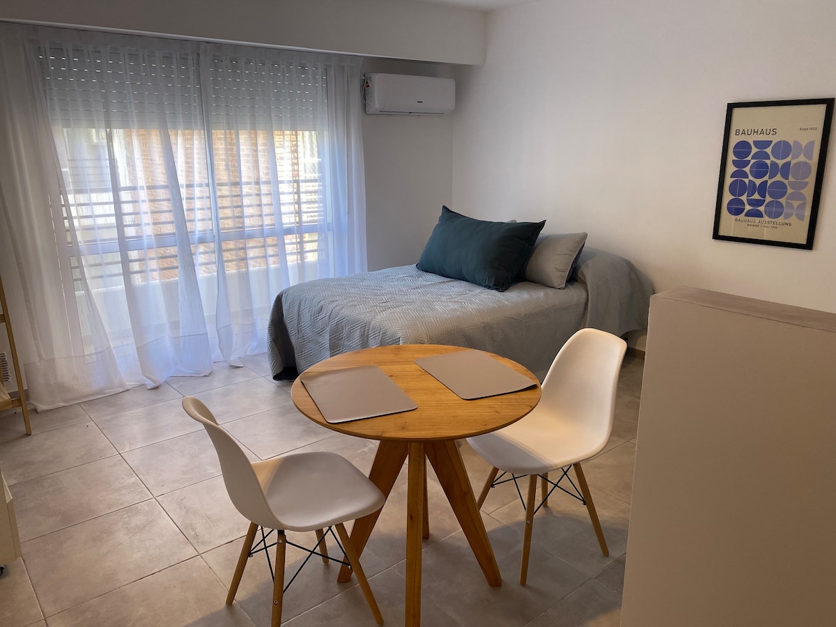 A cozy room features a spacious bed dressed in a light blue coverlet with dark pillows. A round wooden table with two white chairs is positioned in front of a window adorned with sheer curtains, allowing natural light to fill the space.