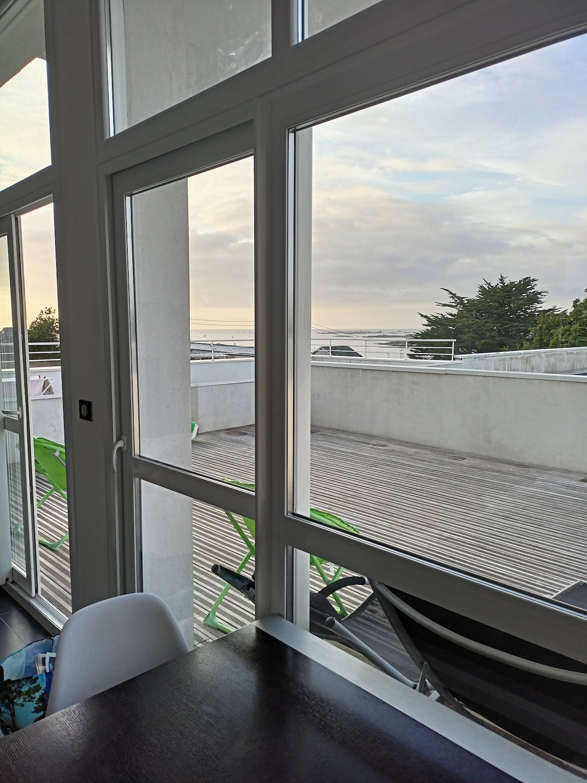 Large windows provide a clear view of the outdoor terrace and ocean beyond. The terrace features wooden flooring and is furnished with green and black lounge chairs, enhancing the connection between indoor and outdoor spaces.