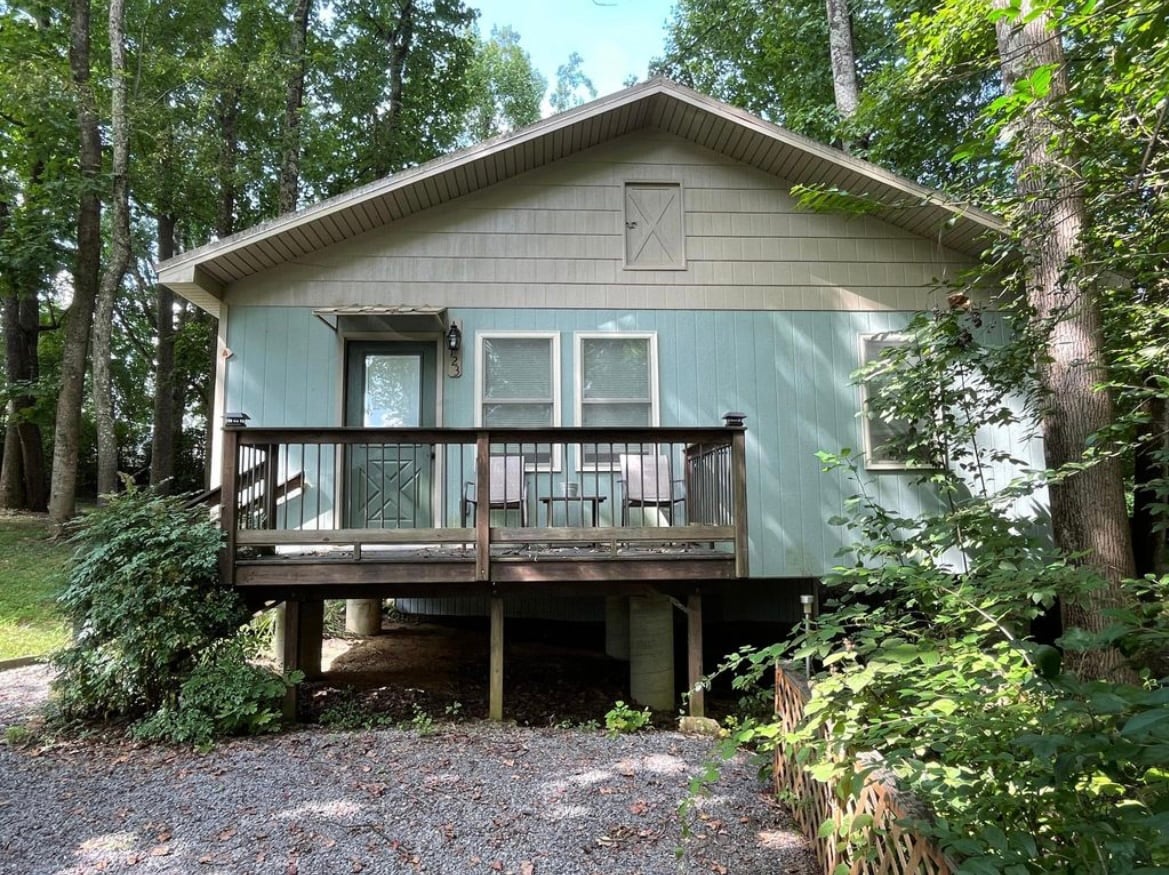 A charming cabin is nestled among trees, featuring a welcoming porch with patio chairs. The light blue exterior contrasts with the surrounding greenery. Gravel leads to the entrance, enhancing the natural setting. Large windows allow sunlight to brighten the inviting atmosphere.