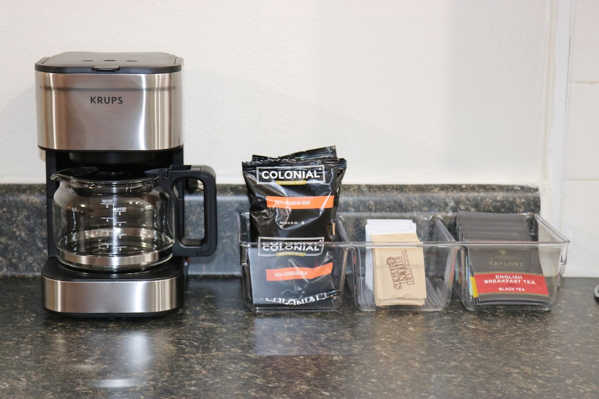 A coffee maker sits on a black countertop, accompanied by neatly organized containers. One container holds coffee packets, while another contains a variety of tea bags and sugar packets. The setup provides guests with convenient options for morning beverages.