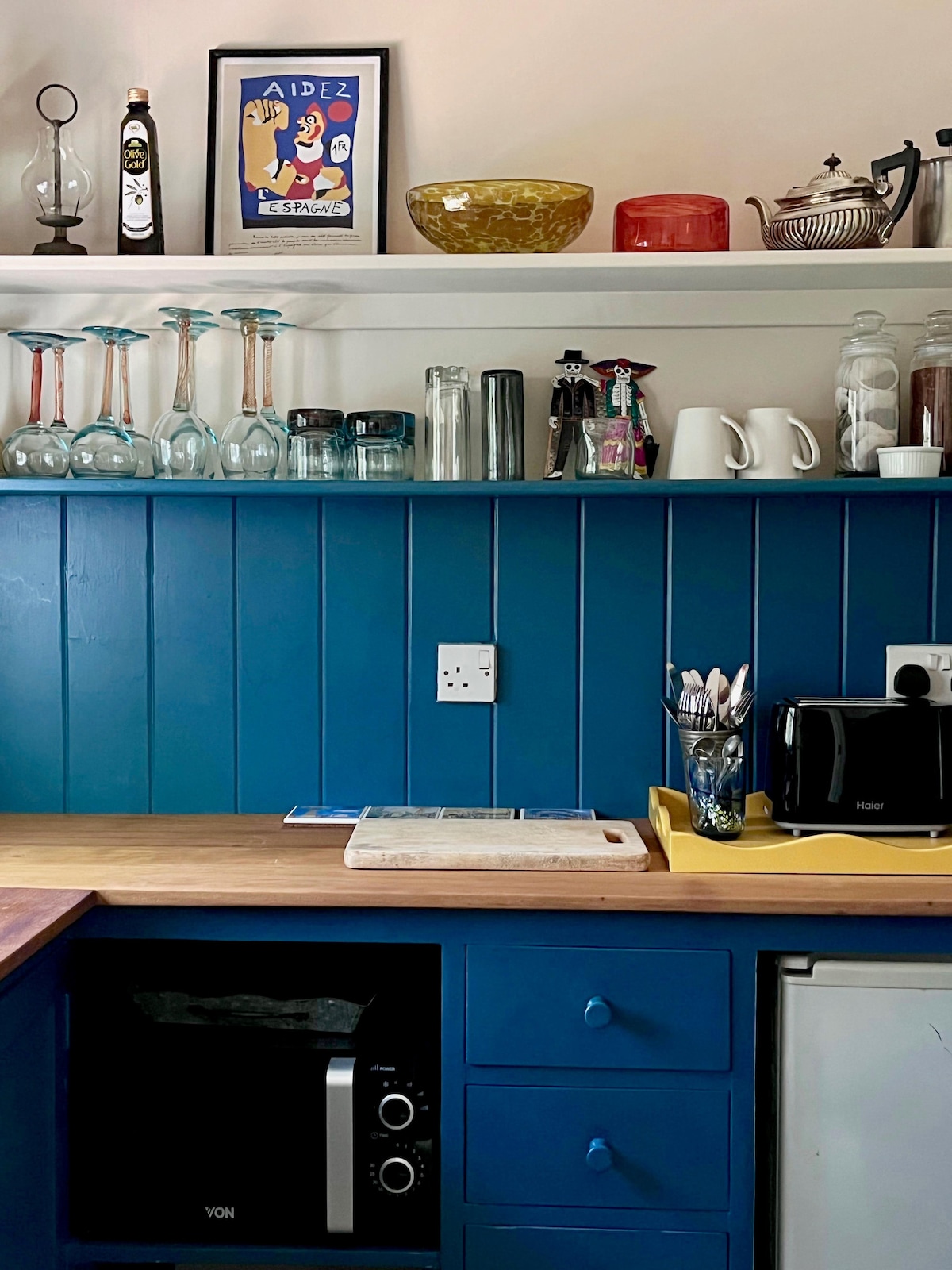 A compact kitchen area features a blue-paneled wall and a wooden countertop. Glassware, cutlery, and a tea set are arranged on a white shelf above. A microwave and a refrigerator are situated below, providing essential appliances for guests.