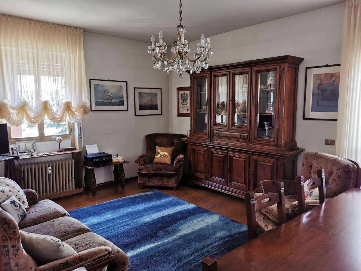 A spacious living room showcases a classic wooden cabinet with glass doors, complemented by a plush, patterned sofa and an armchair. A dining table occupies part of the room, while a soft blue rug adds comfort underfoot. Large windows allow natural light to fill the space.