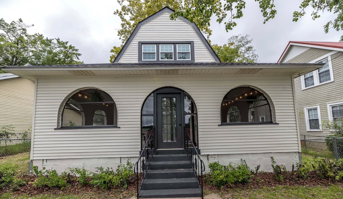 The exterior of the historic home is illustrated, featuring a welcoming entrance with a staircase leading to double doors. Arched windows are present on either side, offering a glimpse of the design's character. Surrounding greenery complements the well-kept facade.