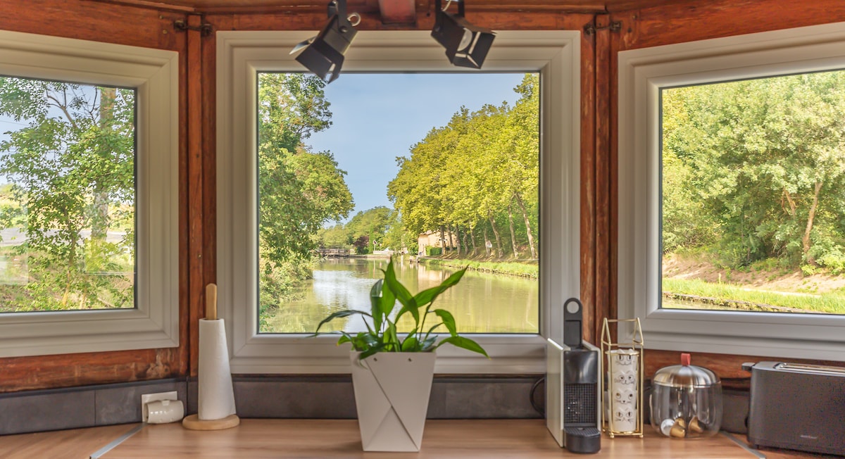 Large windows provide serene views of the canal and lush greenery outside. A decorative plant sits on a wooden countertop, enhancing the space. Natural light floods the area, creating a bright and inviting atmosphere.