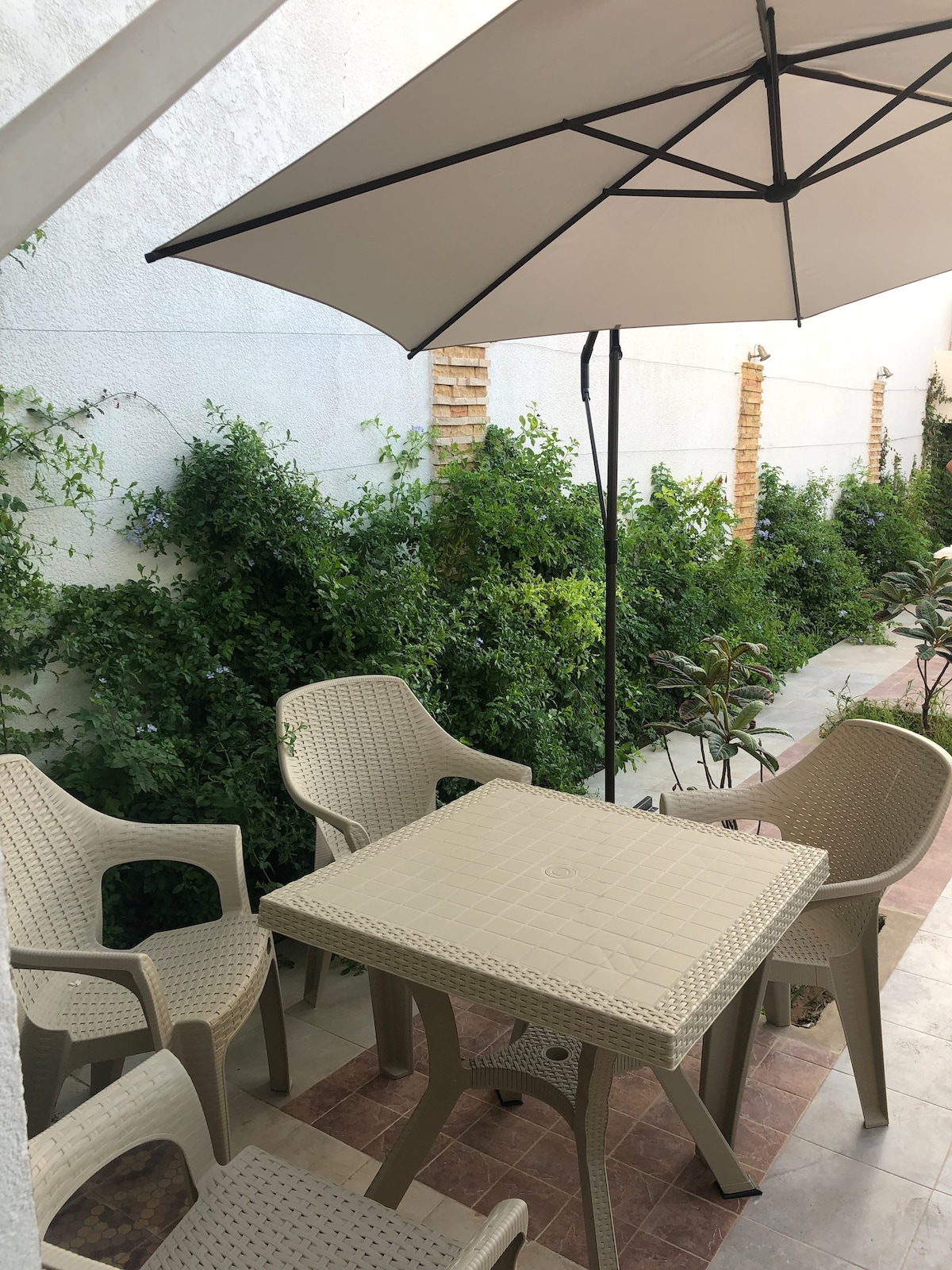 A shaded outdoor seating area is presented, featuring a square table surrounded by four structured chairs. Lush greenery lines the edges, providing a natural backdrop. An umbrella offers shade, creating a tranquil space for relaxation.