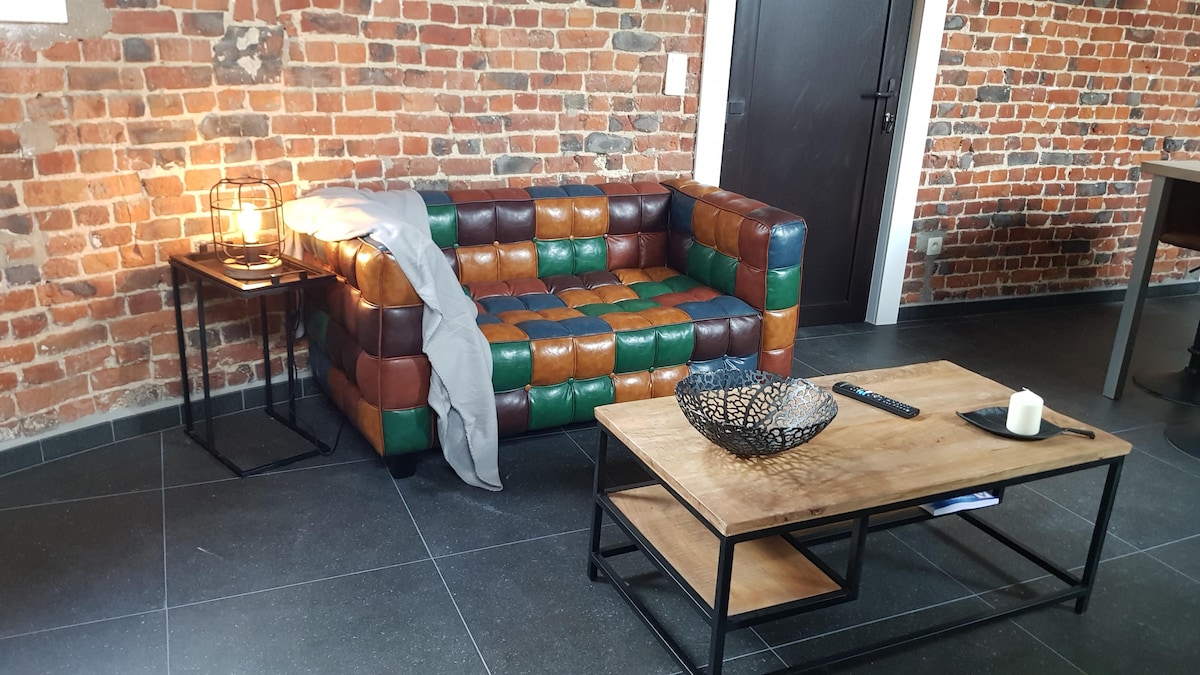 A unique multicolored leather couch is positioned against a brick wall, accompanied by a wooden coffee table. A decorative bowl sits on the table, and a soft throw is draped over the couch. Warm light emanates from a lantern placed on a side table.