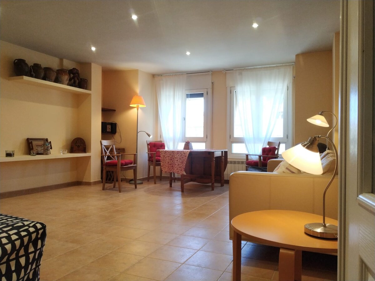 A spacious living area features natural light flowing through large windows with sheer curtains. A light-colored sofa and wooden coffee table are present, along with a dining table surrounded by four chairs. Decorative pottery adds character to the shelves.