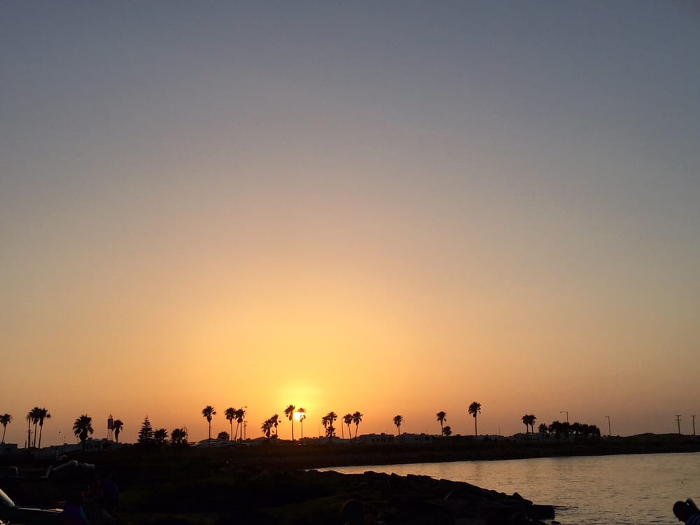 A sunset scene is captured over a tranquil body of water, with silhouettes of palm trees lining the horizon. The sky transitions from orange to soft purple, creating a serene atmosphere as daylight fades.