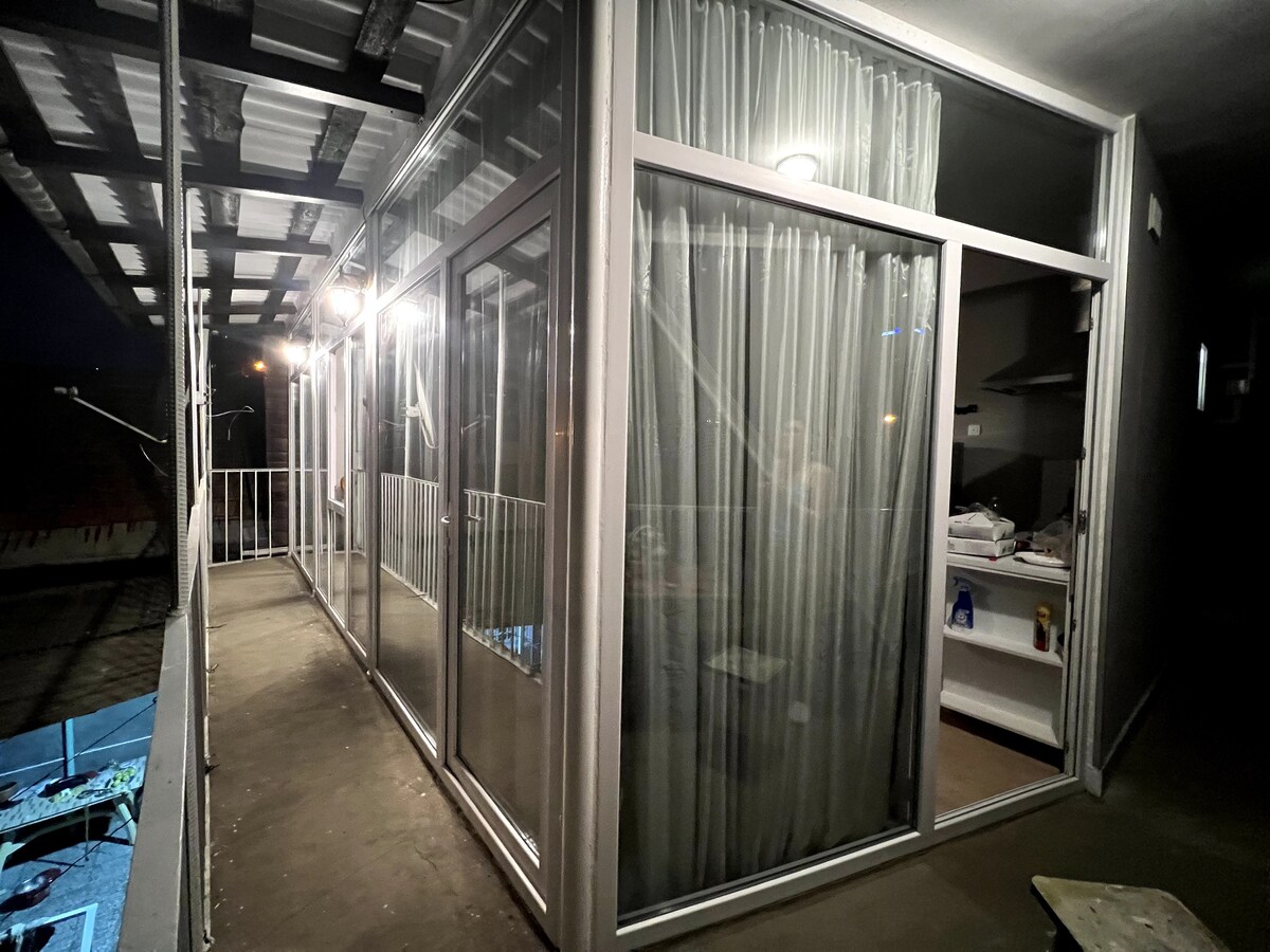 A balcony area is shown with large glass panels providing views of the courtyard below. Sheer curtains are gently flowing, and soft lighting is displayed. The separation between indoor and outdoor spaces is emphasized by the sliding doors leading to the living area.