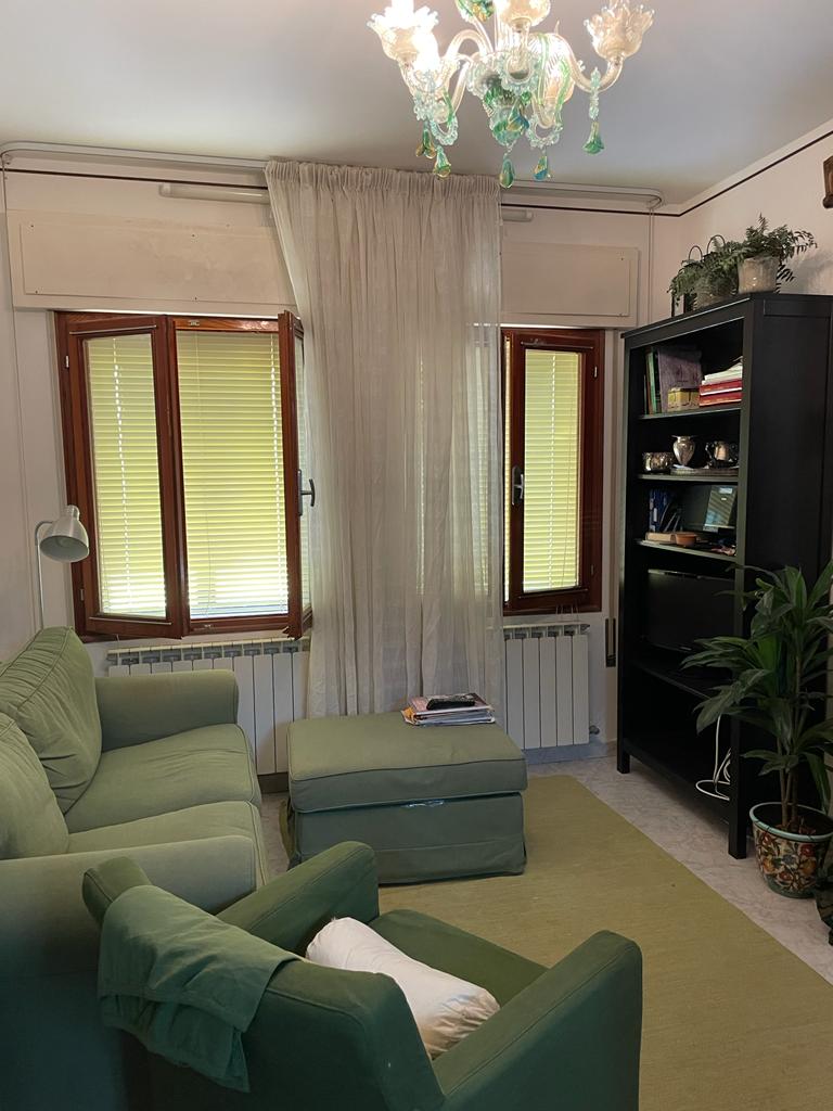 A cozy living area is presented, featuring a green sofa and an ottoman. Natural light filters through double windows adorned with sheer curtains. A tall black bookshelf and a potted plant add character, while a light fixture hangs from the ceiling, providing a gentle ambiance.