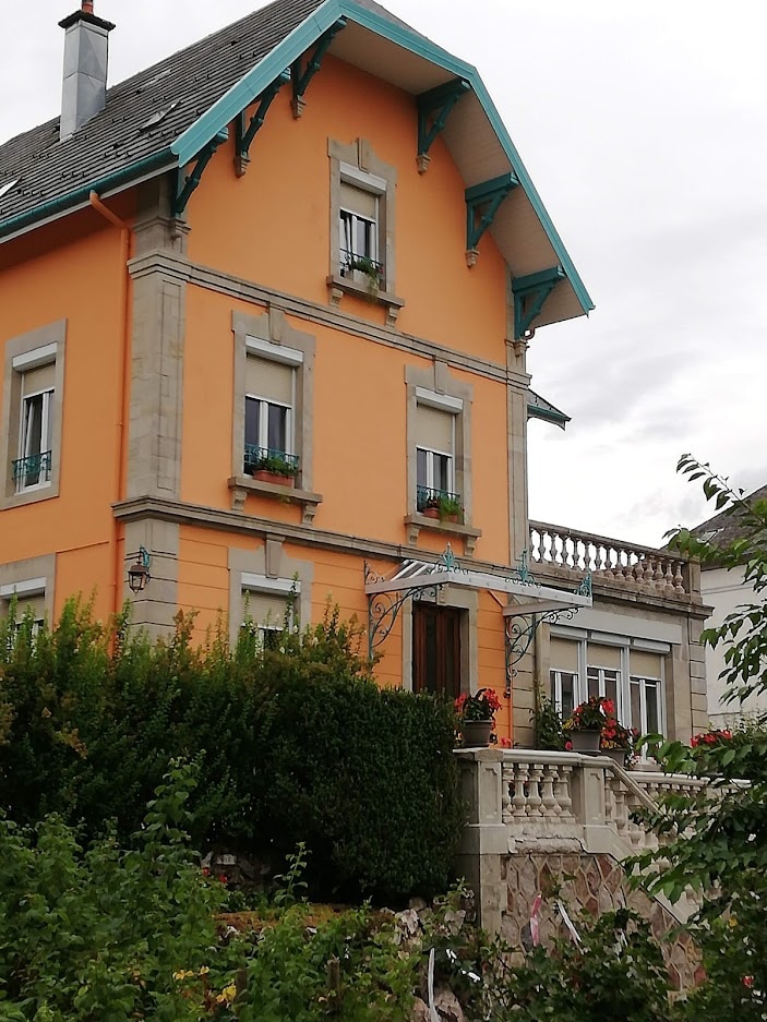 A charming three-story house in vibrant orange features decorative wooden eaves and flower boxes under the windows. A stone balcony is visible on the second level, and lush greenery surrounds the base, adding to the inviting exterior.