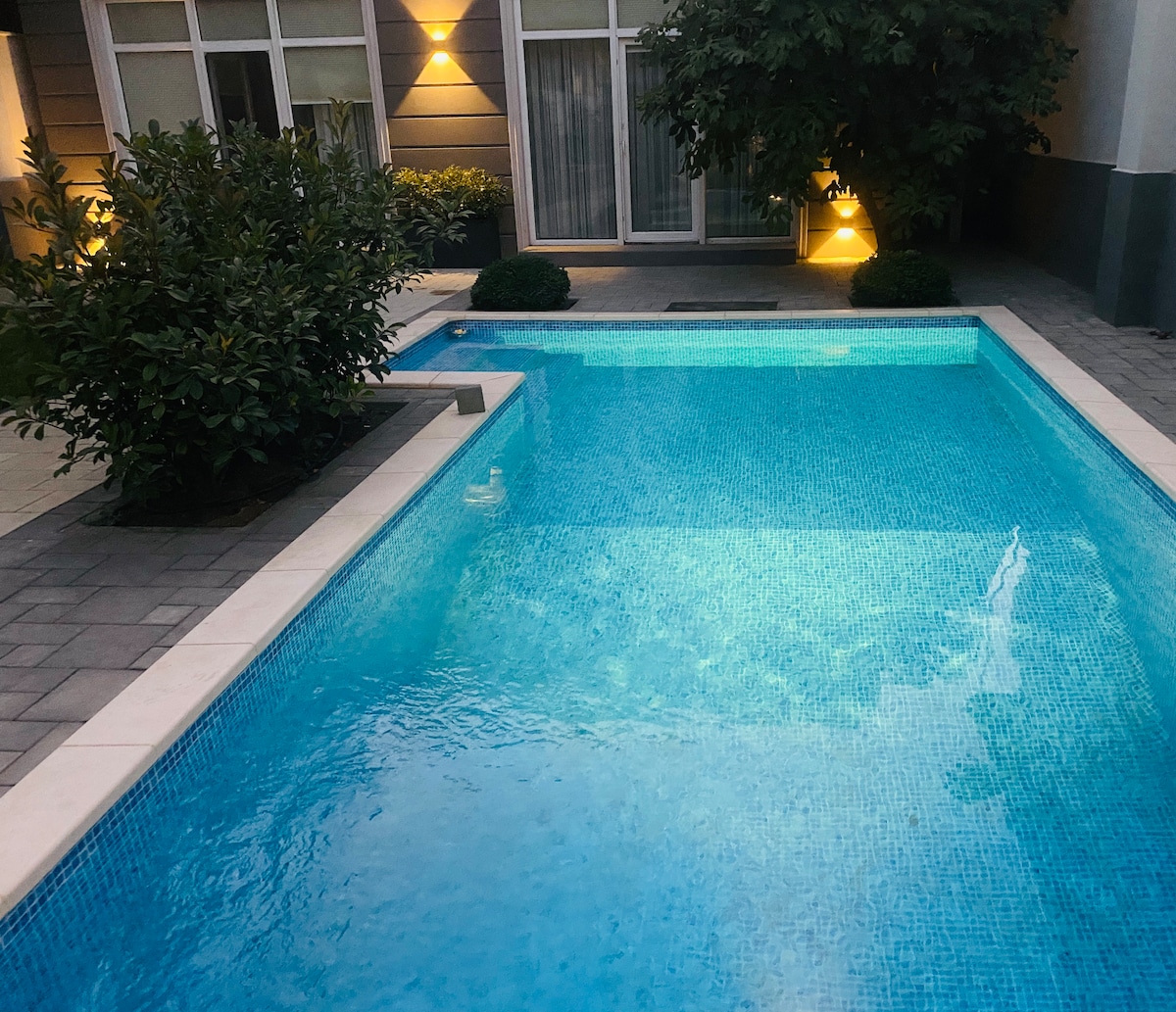 An inviting swimming pool is surrounded by neatly trimmed greenery and soft lighting. The clear water reflects the surroundings, enhancing the relaxing ambiance of the outdoor area. The pool is framed by a cool-toned stone patio, providing ample space for lounging.
