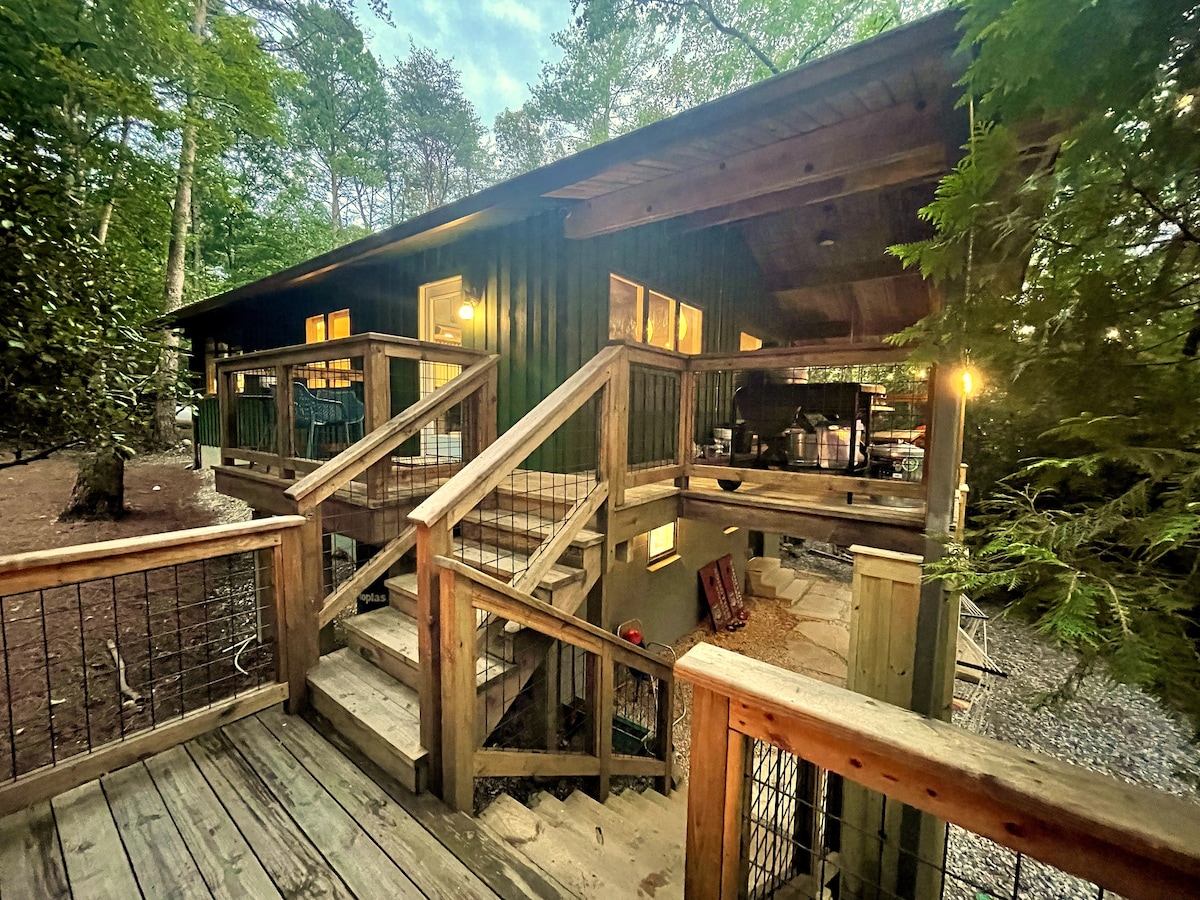 The exterior entrance to the cabin is framed by lush trees, featuring wooden steps leading up to a spacious deck. Soft lighting illuminates the area, highlighting the inviting outdoor seating and the rustic charm of the cabin's design.