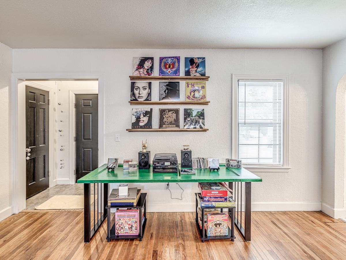 A vibrant green table displays a record player and speakers, surrounded by various vinyl records and music-related books. A wall-mounted shelf showcases colorful album covers. Natural light enters through a window, illuminating the wooden floor and enhancing the room's inviting atmosphere.