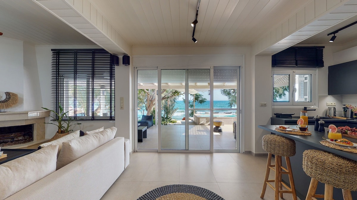 An open living area is featured with a beige sofa facing large sliding glass doors that provide a view of the private beach and sea. A small dining table is set with refreshments, complementing the modern kitchen space in the background.