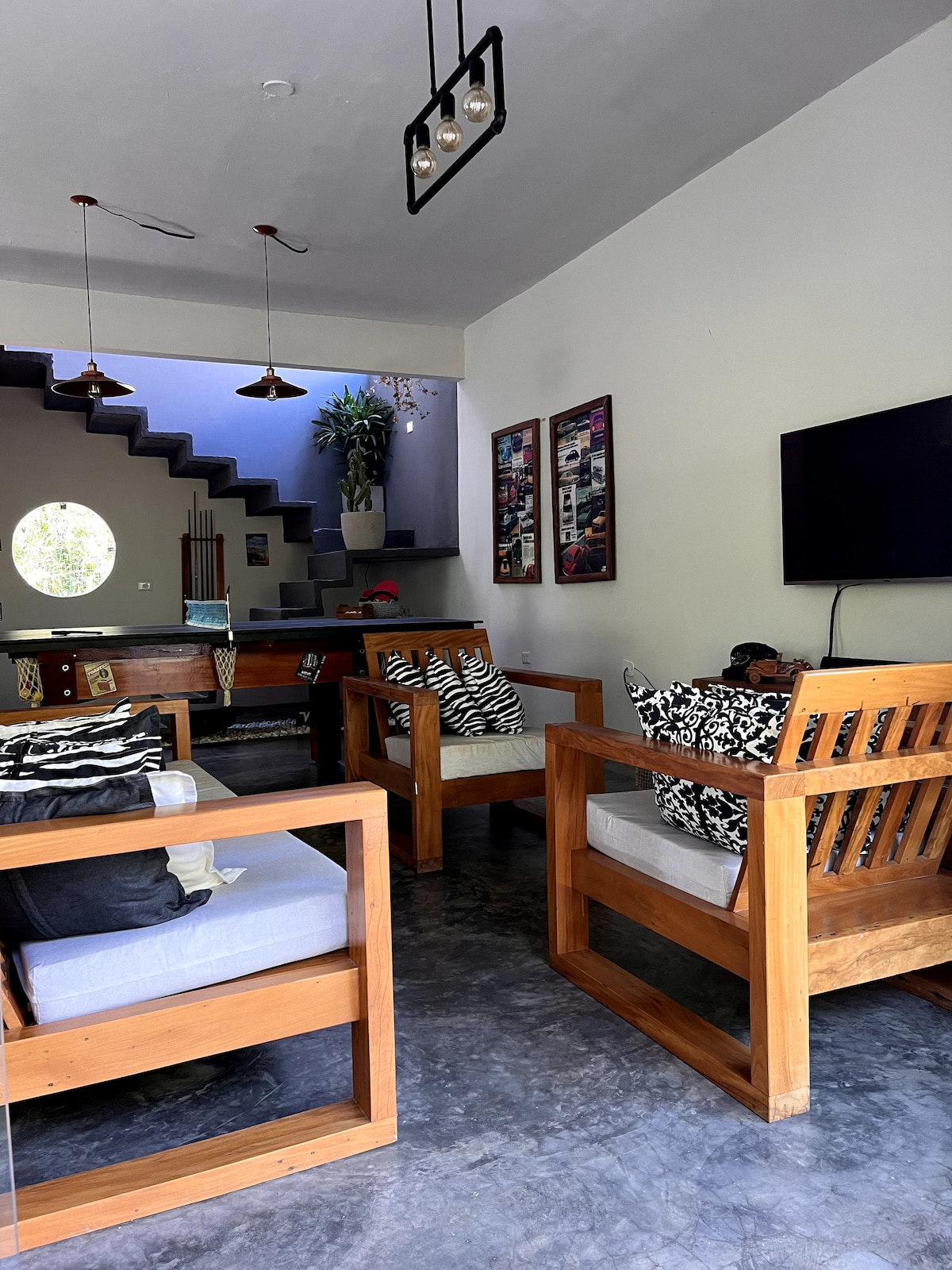 A comfortable living area features wooden armchairs adorned with patterned cushions. A television is mounted on the wall, while a staircase leads to an upper level. Natural light enters through a circular window, enhancing the open layout.