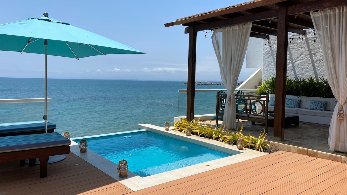 A serene outdoor space features a pool seamlessly merging with the ocean view. An umbrella shades a lounge area, complemented by cozy seating under a wooden pergola. Lush greenery enhances the atmosphere, creating an inviting space for relaxation by the water.