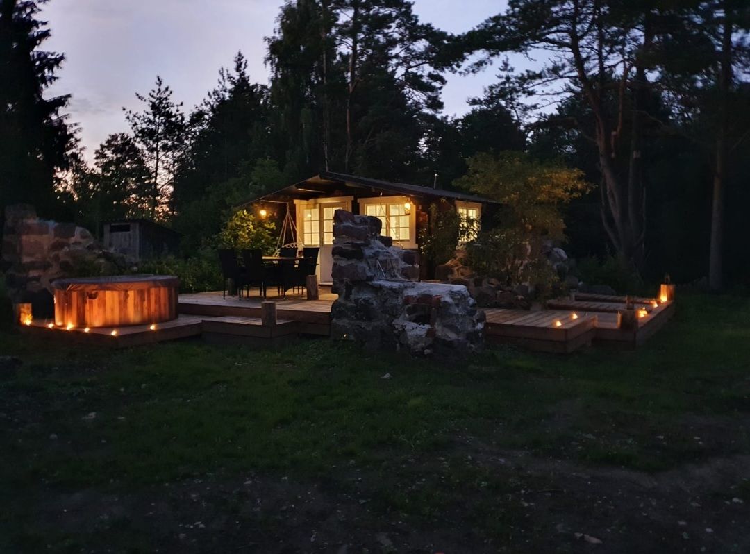 A rustic cabin is softly illuminated at dusk, surrounded by nature. Wooden decking features seating arrangements, and a hot tub is positioned nearby. Stone pathways are lined with glowing lanterns, creating a serene atmosphere in the outdoor space.