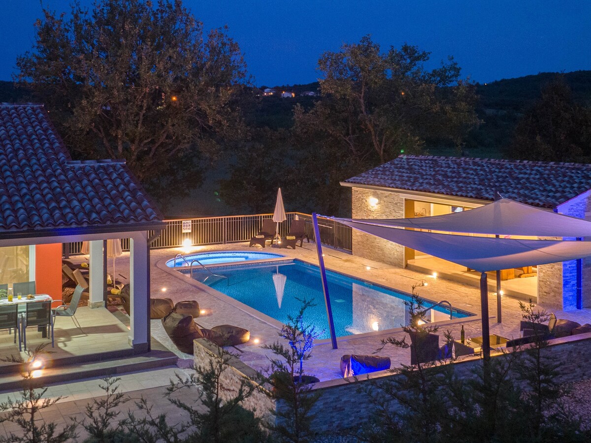 An aerial view of a well-lit outdoor area features a spacious pool illuminated in various colors, surrounded by an inviting patio. Nearby, a covered terrace provides dining space. A stylish garden is visible, adding to the tranquil evening ambiance.