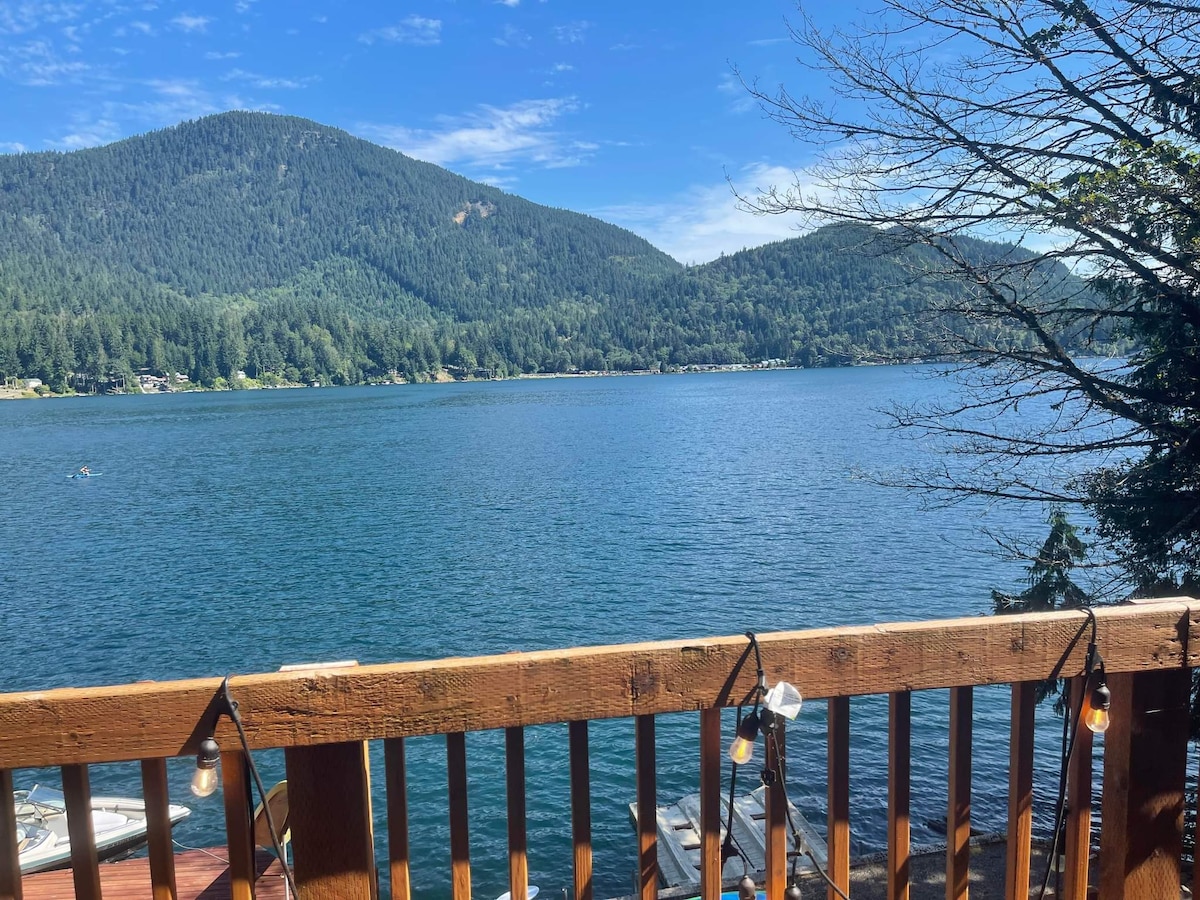 A serene view of Lake Sutherland is presented from a wooden deck. Gentle waves ripple across the water, reflecting the surrounding green hills and blue sky. Mountain peaks rise in the distance, completing the idyllic lakeside scenery.