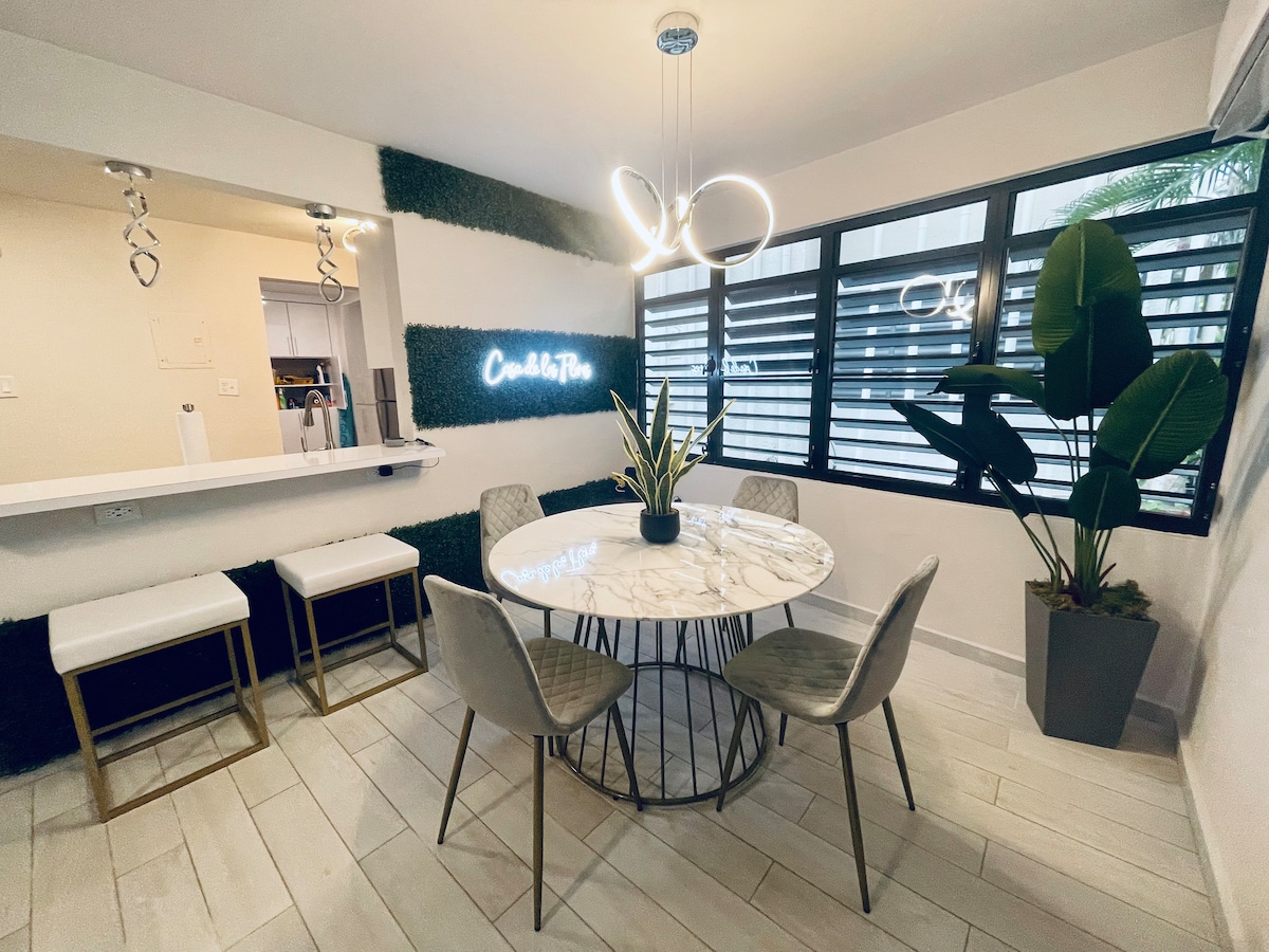 A dining area featuring a round marble table surrounded by four upholstered chairs is visible. Two bar stools are positioned near a counter. Large windows with horizontal slats allow natural light to brighten the space, complemented by a modern light fixture above the table.