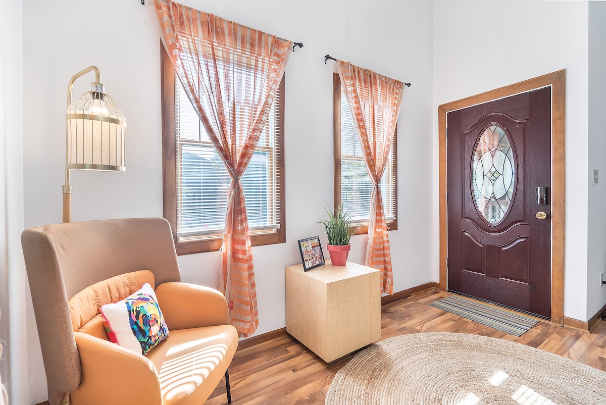 An inviting entryway features warm wooden floors and large windows adorned with orange curtains. A comfortable armchair sits beside a small, light-colored table that holds a potted plant. The front door, with a decorative glass panel, greets guests with a warm welcome.