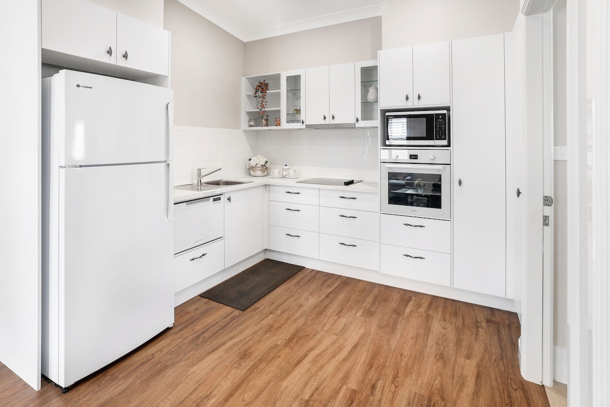 A modern kitchen features white cabinetry and appliances, including a refrigerator, oven, and microwave. A streamlined countertop provides ample workspace, and light wood flooring adds warmth to the area. Decorative elements are arranged on the shelves, enhancing the overall design.