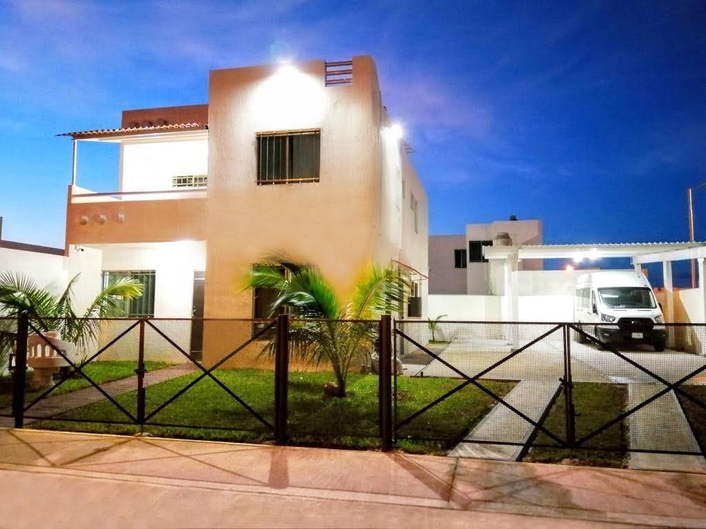 The two-story house is illuminated against the evening sky, displaying a modern design. A neatly landscaped front yard features palm plants. A driveway is visible, accommodating a vehicle under a covered area. The entrance is marked by a secure fence and gate.