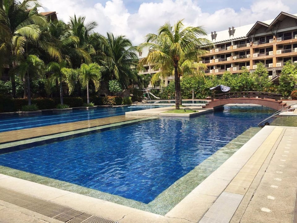 An expansive swimming pool is surrounded by lush palm trees, reflecting the bright blue sky. A wooden bridge crosses over a smaller section of the pool, blending seamlessly with the tropical landscape. The modern condominium building is visible in the background, enhancing the resort-like environment.