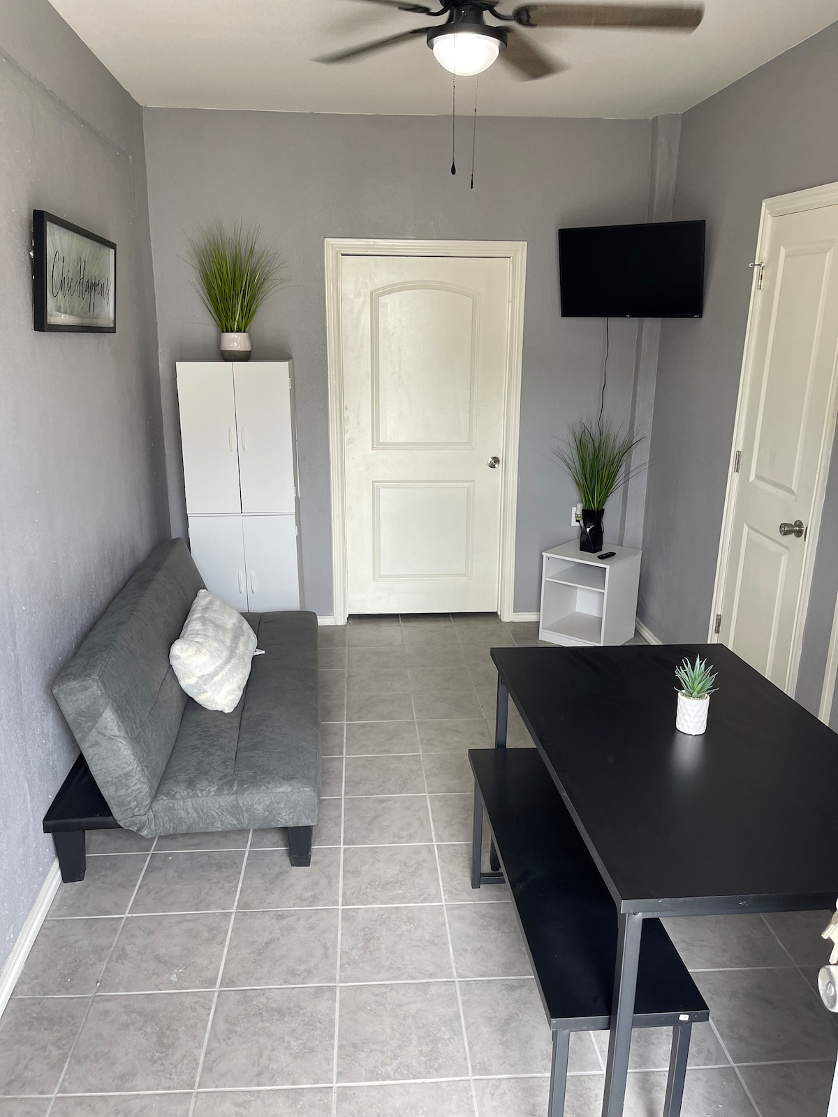 A compact living space features a dark futon and a small black dining table with two chairs. Multi-colored plants adorn the surfaces, while a flat-screen TV is mounted on the wall. Light gray tiles create a clean and bright atmosphere.