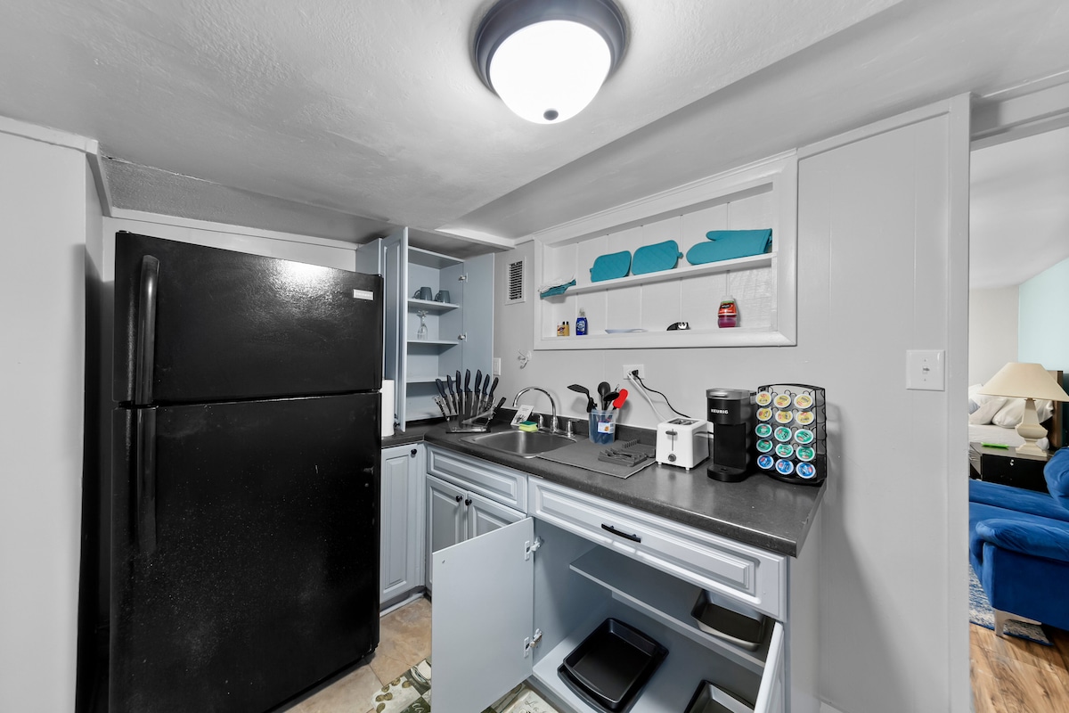 A well-equipped kitchen area features a black refrigerator, grey cabinetry, and a countertop with essential cookware. Shelves hold colorful kitchen items, while the cabinets below provide additional storage options. Natural light from a nearby window brightens the space.