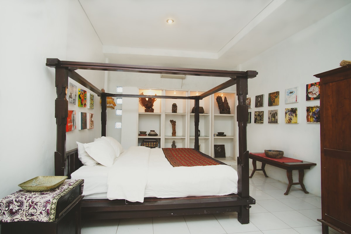 A spacious bedroom features a four-poster bed with crisp white linens and decorative pillows. The walls are adorned with various artworks, complemented by wooden shelves displaying artifacts. A wooden cabinet and a low table add to the room's practicality and charm.