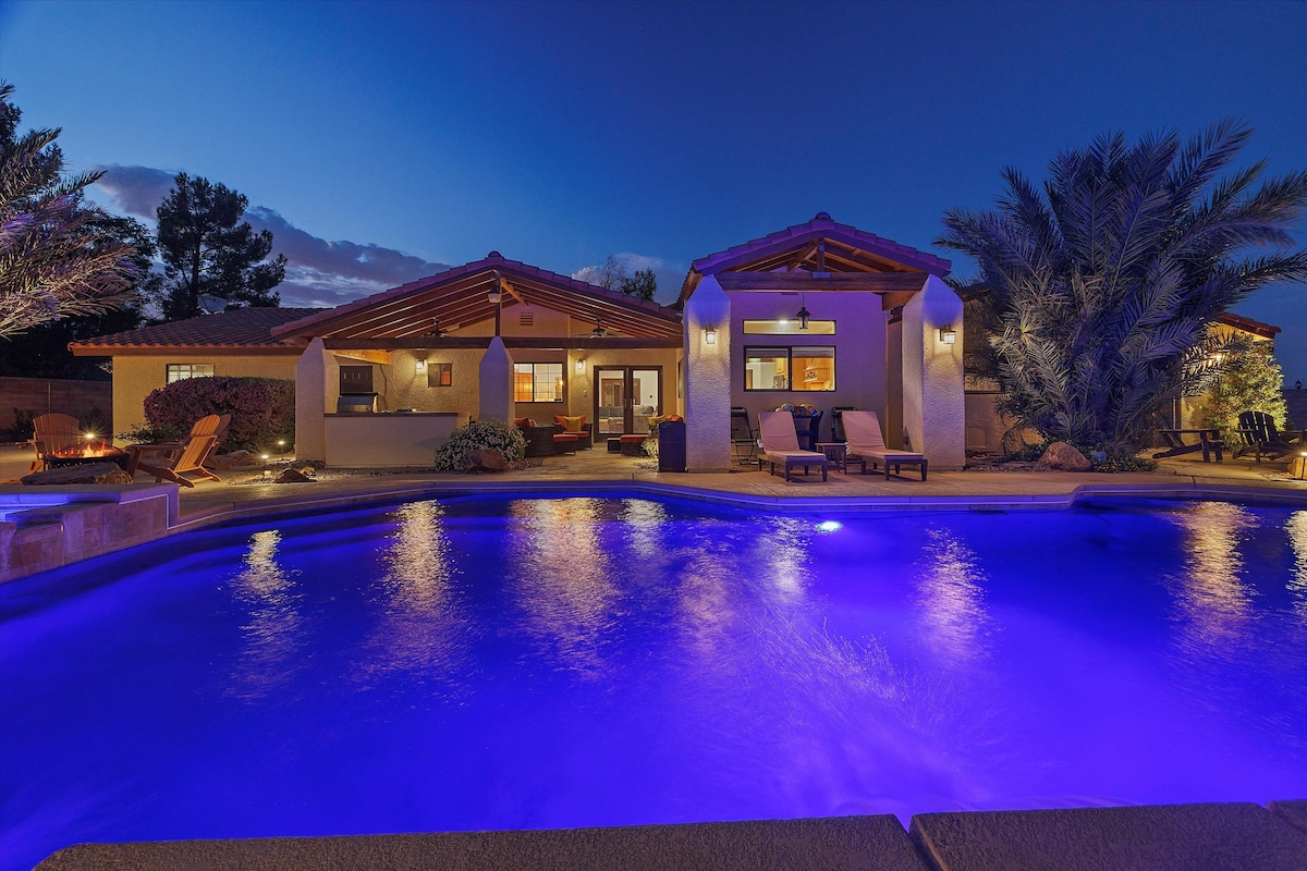 An inviting outdoor space features a heated pool with soft purple lighting, surrounded by well-maintained landscaping. The single-story home showcases a patio with lounge chairs and access to an outdoor kitchen, emphasizing a serene atmosphere at dusk.